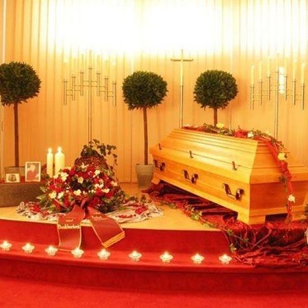 Hölzerner Sarg mit weiß roter Blumendekoration und in gleichen Farben gehaltenem Kranz in einer hell erleuchteten Kapelle