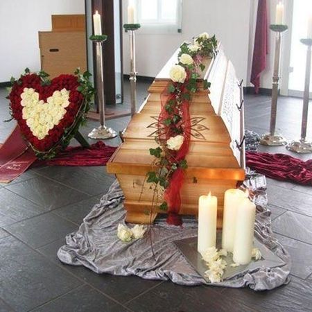 Hölzerner Sarg mit gesteckten, weißen Rosen auf einer roten Schärpe sowie beigestelltem gesteckten Blumenherz in weiß und rot wie auch weißen Kerzen in hohen Kerzenständern
