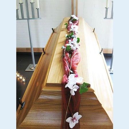 Sarg mit gesteckten, weißen Blumen und grünem Blattwerk auf einer roten Schärpe