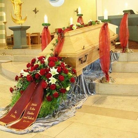 Sarg mit bestickter, roter Schärpe und üppigem Blumenkranz mit weißen und roten Blüten sowie bestickter Schärpe in passender Farbe gehalten