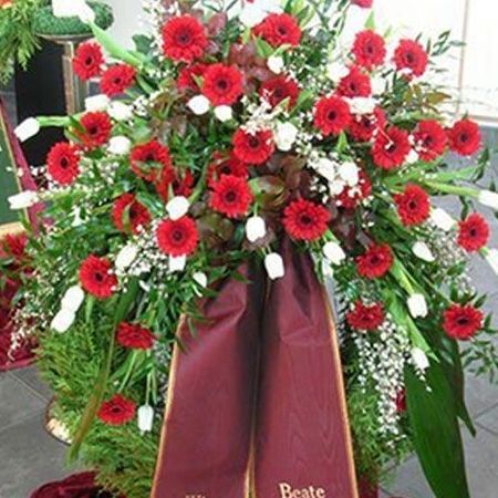 Üppiger Trauerkranz bestehend aus roten und weißen Blumen, grünem Beiwerk und roter, bestickter Schärpe