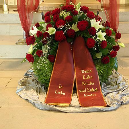 Üppiger Trauerkranz bestehend aus roten und weißen Blumen, grünem Beiwerk und roter, bestickter Schärpe