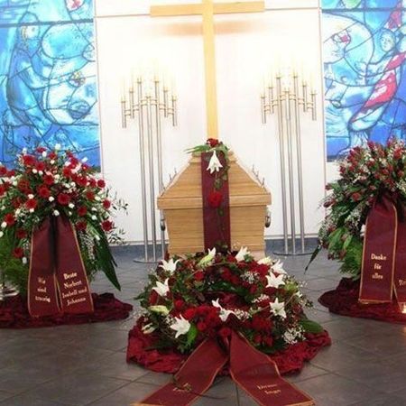 Üppige Trauerkränze bestehend aus roten und weißen Blumen, grünem Beiwerk und roten, bestickten Schärpen seitlich und vor einem Sarg drapiert
