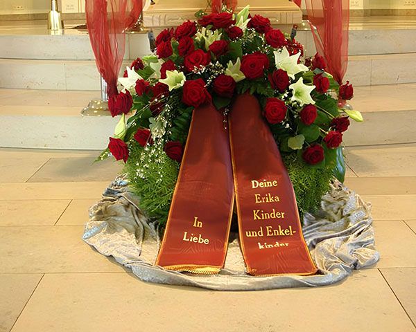 Trauerkranz bestehend aus roten und weißen Blumen, grünem Beiwerk und roter, bestickter Schärpe
